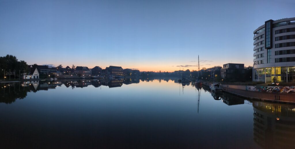 View from the middle of the Leer harbour bridge at sunrise: the colours of the reflection of the sky in the water from where you stand fade from black into dark blue, light blue and finally the light orange of the dawn. Then the gradient is interrupted by the dark line of buildings framing the harbour, their windows illuminated. Looking further up the gradient repeats in reverse, this time as the sky itself: light orange from the morning sun behind the horizon fading into light blue and dark blue towards the top of the image. The whole scene has vibe of tranquility and peace.