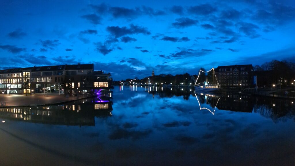 Dark-ish blue sky with a few dark blue clouds reflected in the still water of the harbour, ship with a chain of lightbulbs on the right, on the left illuminated buildings
