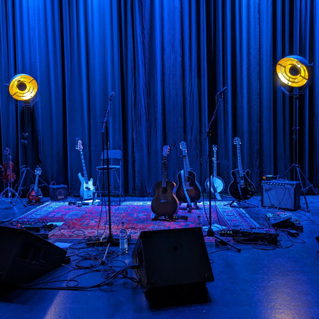 Stage in blue light with two golden spot lights on each side, oriental carpet on the floor, several acoustic guitars and bass standing around