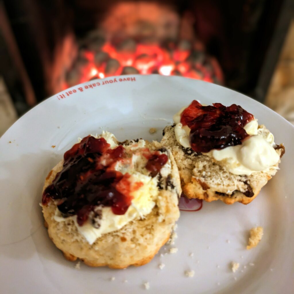 Two half scones with clotted creme and red jam on a white plate in front of a glowing fireplace. On the edge of the plate, upside down from the viewer’s perspective, red cursive letters say ”Have your cake and eat it“