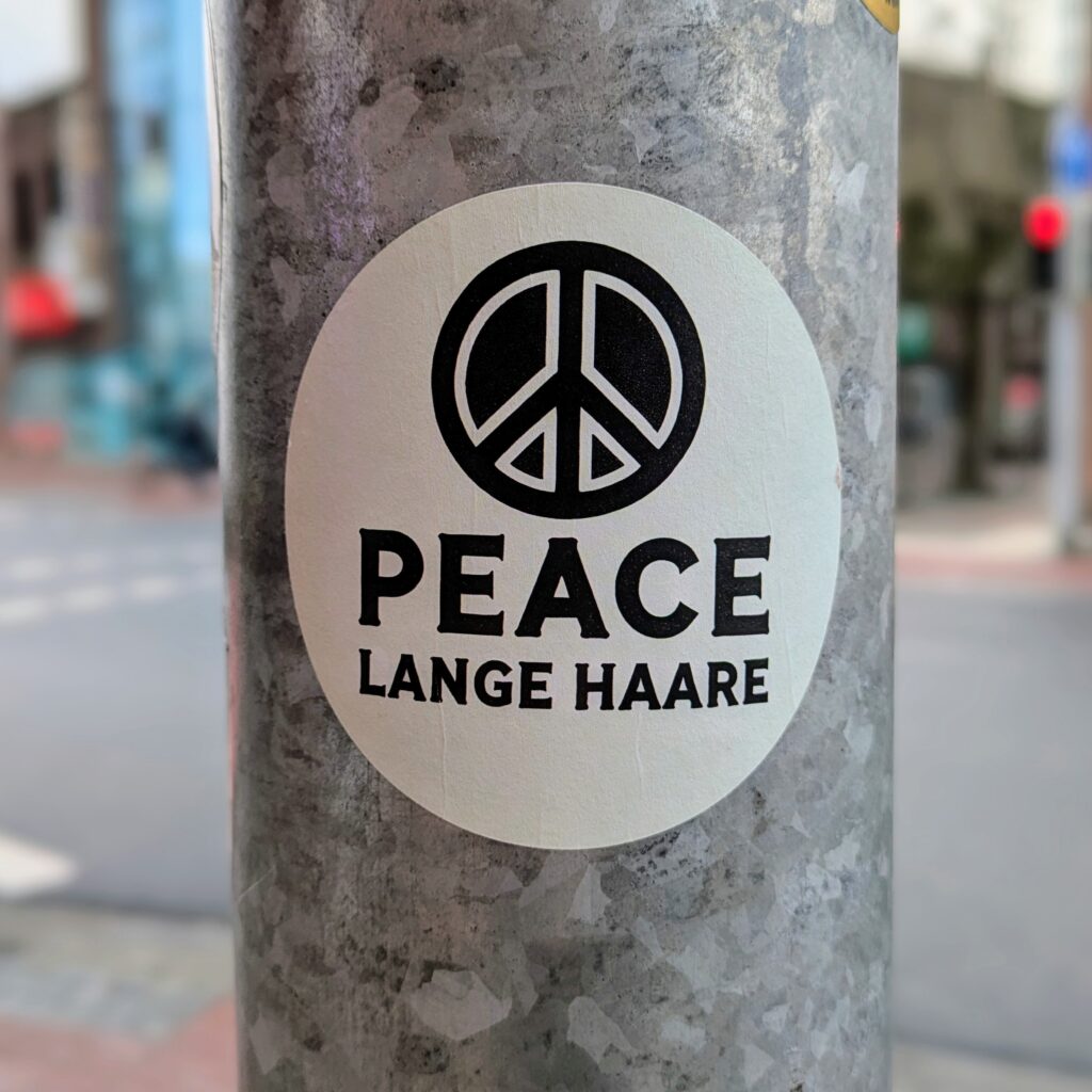 White sticker on a metal traffic light post showing the peace symbol, black caption saying ‘Peace (new line) Lange Haare’ (long hair)