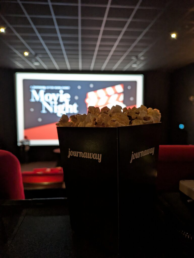 Branded black popcorn bucket with white logo font on a blurred backdrop of a cinema screen showing an event flyer in red and black with white serif font spelling “Movie Night”