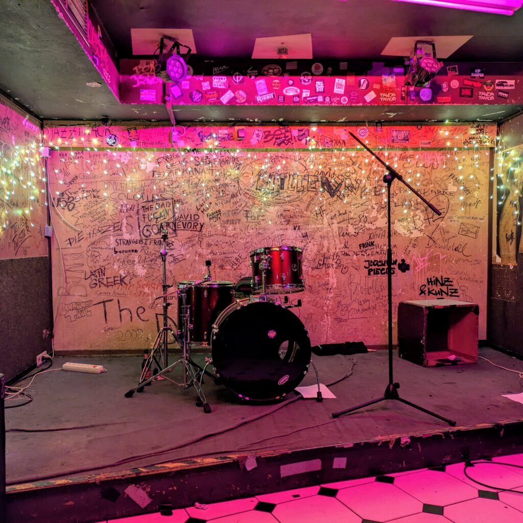 Red drum kit on the empty club stage at Session, Bonn. Purple stage lights on, a wall of band graffiti in the background. Authentic live venue vibe, sweat, cigarette smoke and all 