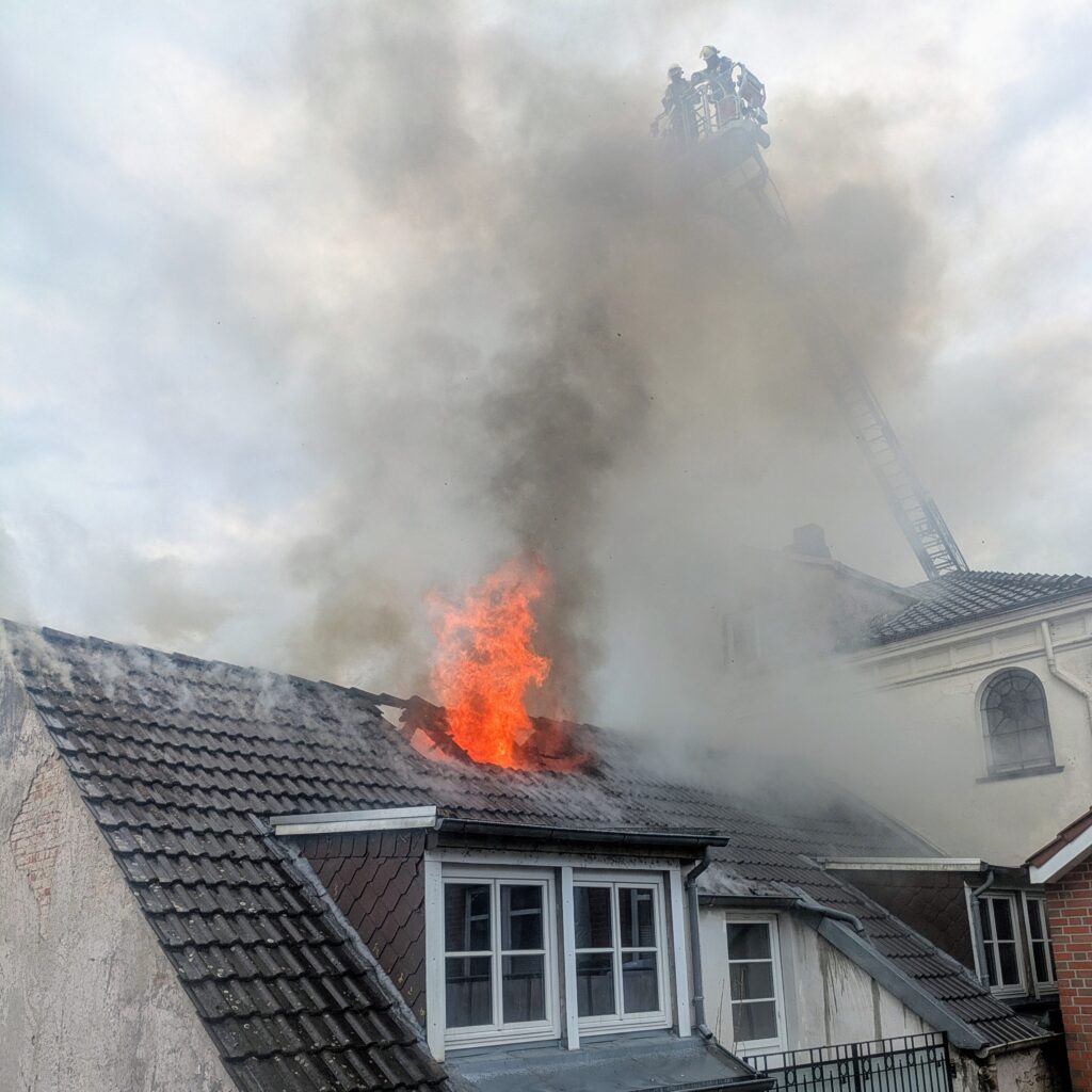 Roof top in flames and smoke, firefighters extinguishing from the top of an extended latter hovering above 
