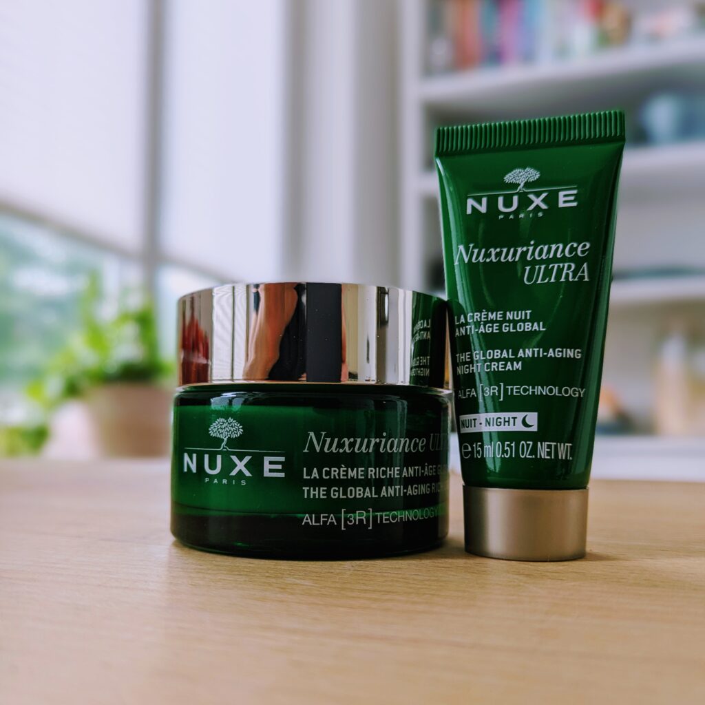 Nuxe face cream jar and tube, both in dark green with white font and lids, on a wooden table.