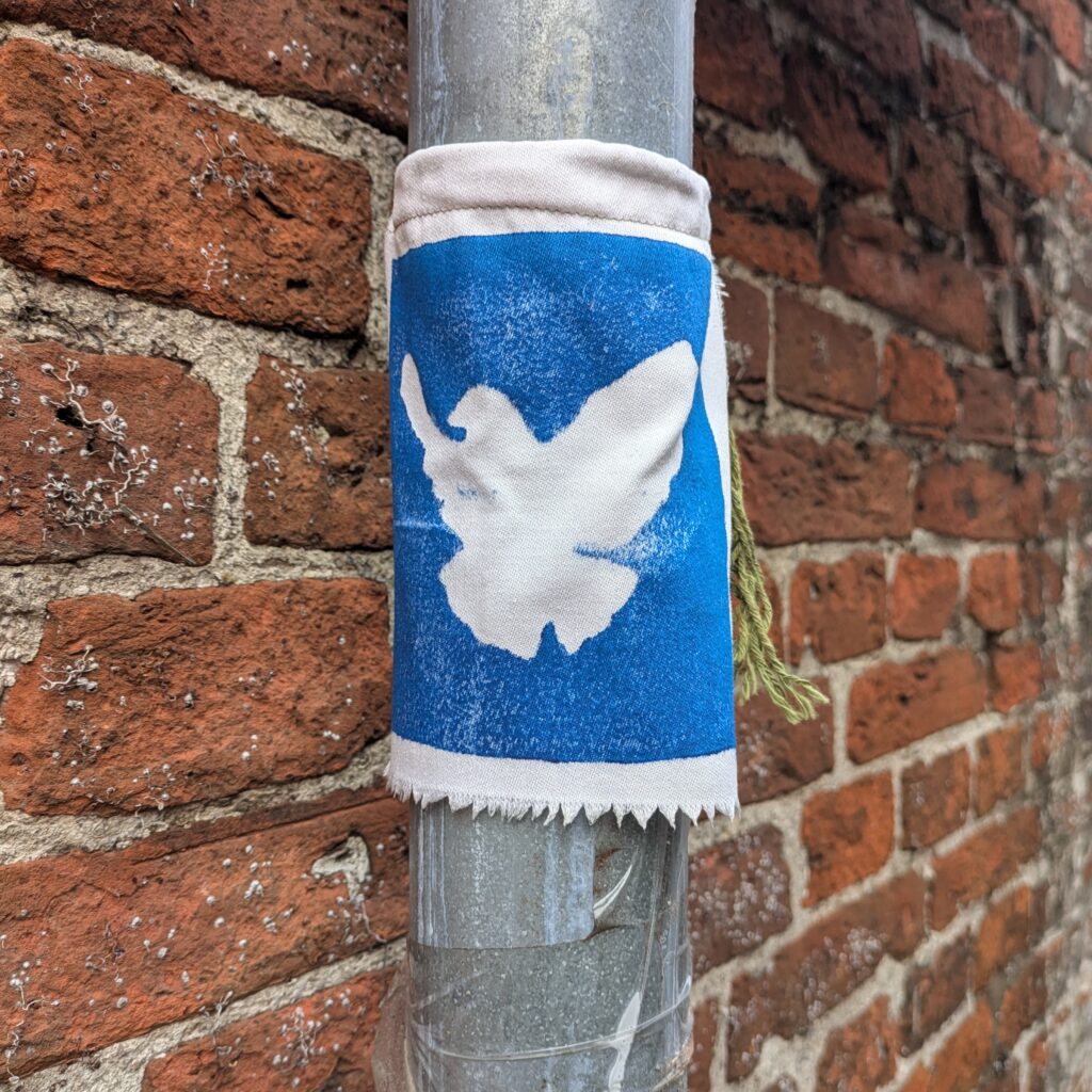 A blue and white fabric band is wrapped around a metal street pole fixed to a red brick wall. The band shows a white, abstract dove silhouette on a blue background. The fabric edges are frayed and a small green tassel hangs from one side.