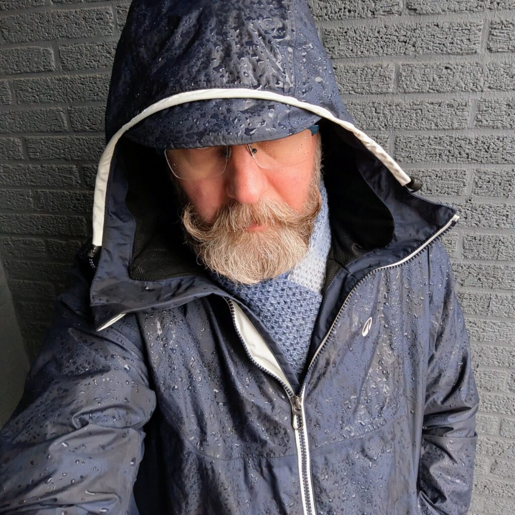 An older man stands against a grey brick wall wearing a navy waterproof jacket with the hood up. Raindrops cover the jacket’s surface. His head is slightly lowered, glasses partly visible under the hood, and he has a thick, neatly groomed beard.