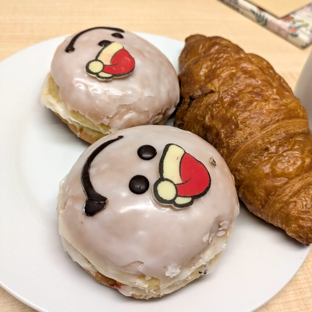 A white plate with two iced Berliners decorated with smiling faces in Santa hats and a croissant beside them on a light wooden table. A stack of napkins on a small plate and a gift wrapped in patterned paper with an envelope lie in the background.