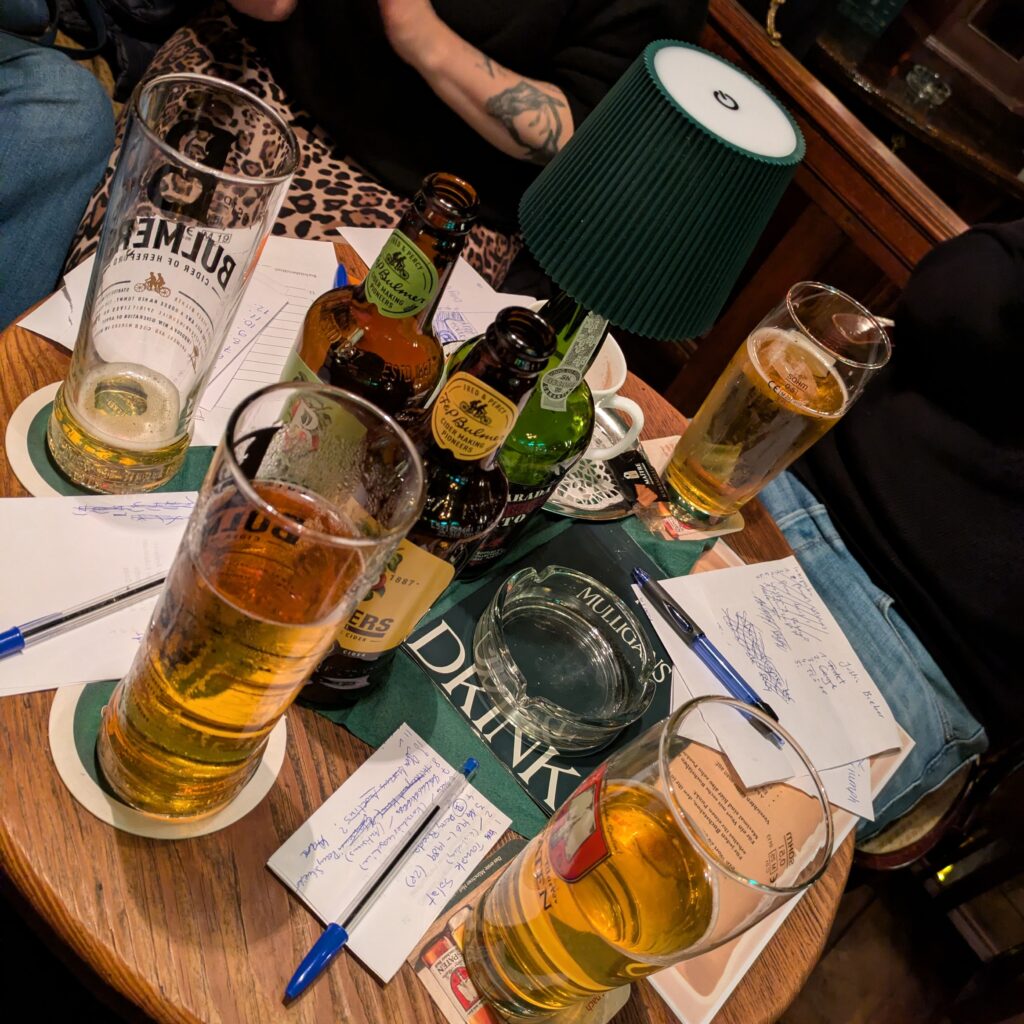 A round wooden pub table crowded with half-full pint glasses of lager and cider, small beer bottles, pens and handwritten answer sheets from a pub quiz. An ashtray sits in the centre alongside a quiz booklet, with people seated closely around the table, their legs and arms visible, suggesting an informal, busy pub setting during a quiz night.