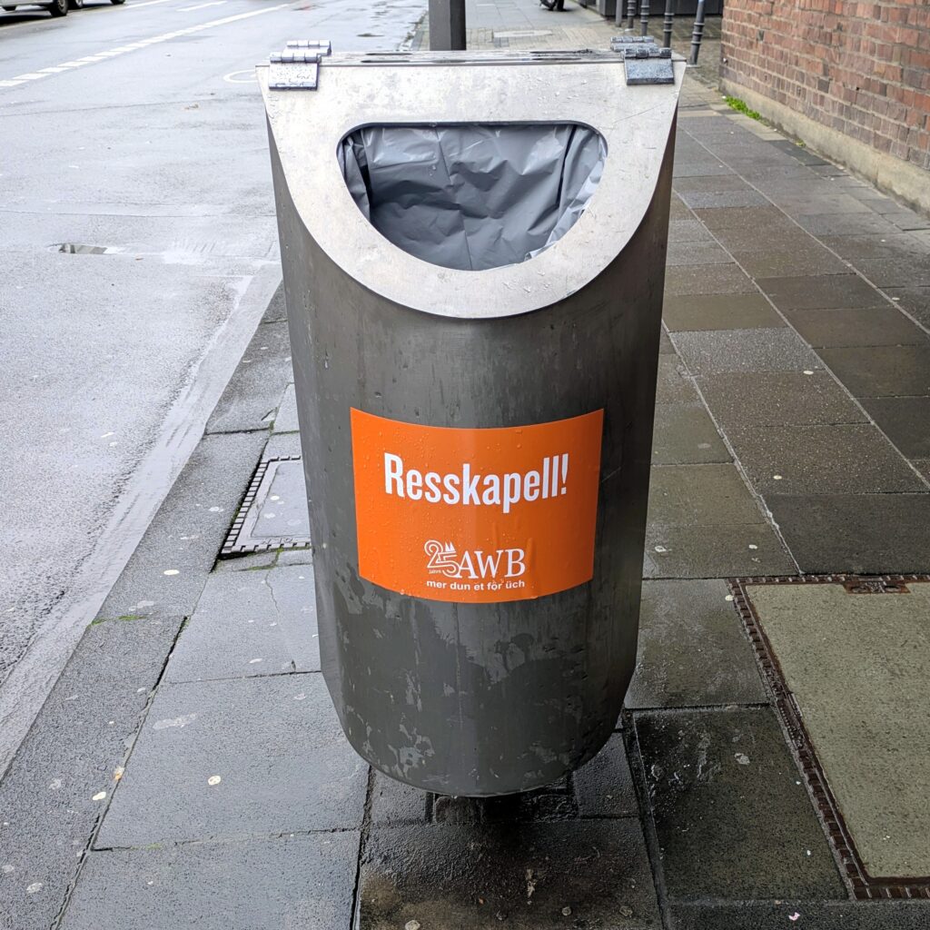 Cylindrical metal rubbish bin on a wet pavement beside a street. The bin has a semi-circular opening lined with a grey bag and an orange sticker reading “Resskapell!” with the AWB logo beneath it. A brick wall runs along the right side and the road with parked cars is visible to the left.