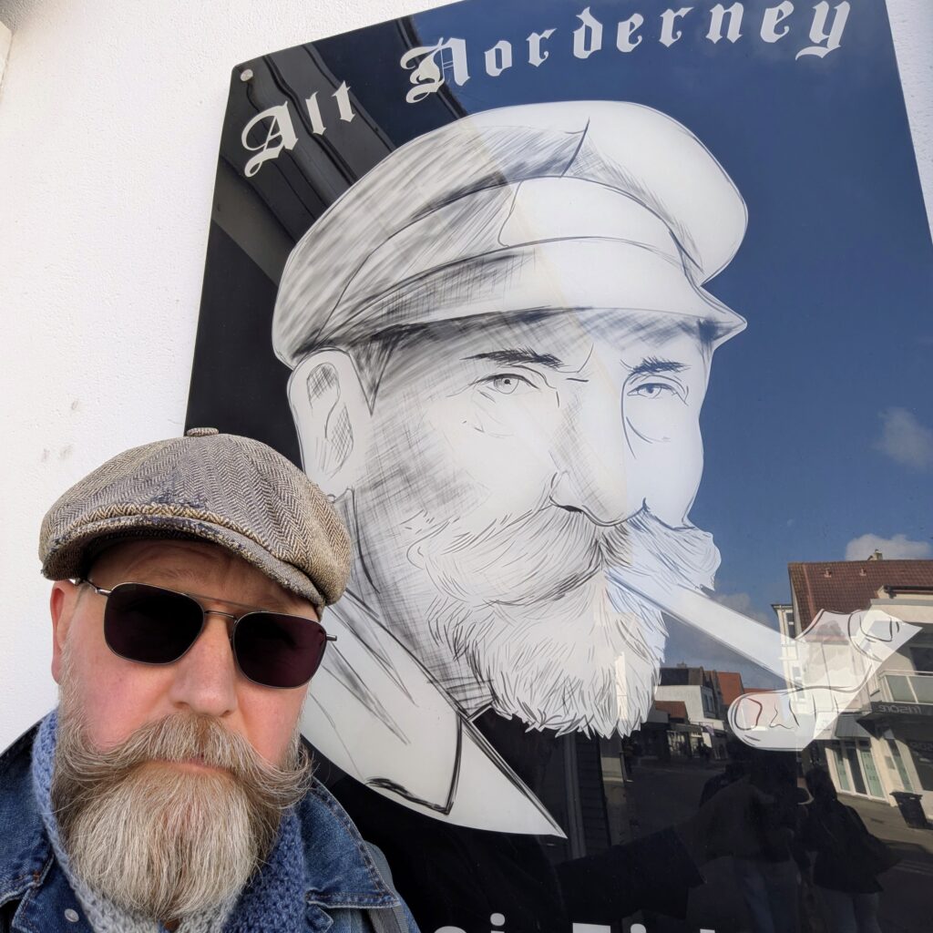 A man with a long grey beard, wearing a tweed flat cap and sunglasses, poses in front of a large sign. The sign features a black-and-white illustration of a bearded man in a captain's hat and the text “Alt Norderney” above the illustration. The background shows an outdoor urban setting with buildings and a clear sky.
